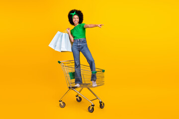 Poster - Full size photo of funny african teen schoolgirl stand pushcart hold bags look direct finger empty space isolated on yellow color background