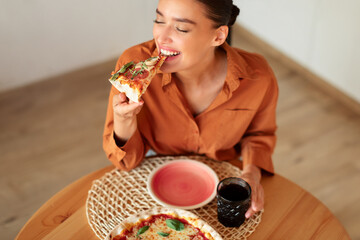 Wall Mural - Excited woman enjoying eating tasty pizza, holding and biting slice, sitting at table in kitchen, free space, above view