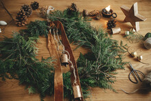 Stylish Christmas Rustic Wreath With Cedar Branches, Ribbon, Vintage Bells, Pine Cones, Twine, Scissors On Wooden Table. Merry Christmas. Winter Holiday Preparations In Atmospheric Room