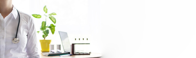 Doctor woman in her office. Doctor with stethoscope on the blured background of her office table with lap top, laboratory dishes, notepads, books and potted flower. Health care concept. Long banner.