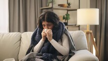 Sick Curly Woman Sitting On Sofa Under The Warm Blanket Coughing Sneezing In Napkin And Having Severe Headache Temperature At Home Unhealthy Female Getting Flu Virus Symptom Cold And Fever Concept
