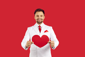 Elegant bearded man in white classic suit with a red tie holding big red heart for valentine's day and smiling isolated on red background in studio, portrait. Valentines day concept, copy space