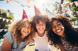Three cheerful teenage friends wearing paper hats celebrating birthday outdoors with colorful confetti and balloons. Teen birthday party in a backyard.