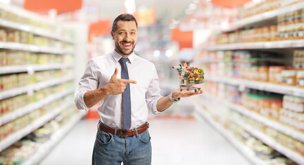 Young smiling man holding a small shopping cart and pointing in a supermarket