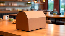 Brown cardboard box for food delivery with a rectangular shape and a triangular lid on a restaurant table. Blurred background with tables, chairs and barware. Food delivery concept.