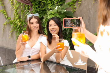 Poster - Beautiful young women with glasses of tasty aperol spritz taking photo in cafe, outdoors