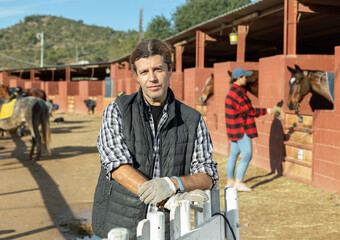 Portrait of positive adult man landowner wearing plaid shirt and posing during daytime on yard of horse stables