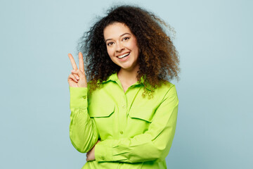 Canvas Print - Young smiling cheerful joyful happy fun latin woman she wear green shirt casual clothes showing v-sign gesture isolated on plain pastel light blue cyan background studio portrait. Lifestyle concept.