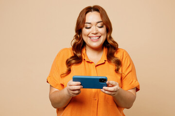 Wall Mural - Young chubby overweight woman wears orange shirt casual clothes use play racing app on mobile cell phone hold gadget smartphone for pc video games isolated on plain beige background studio portrait.