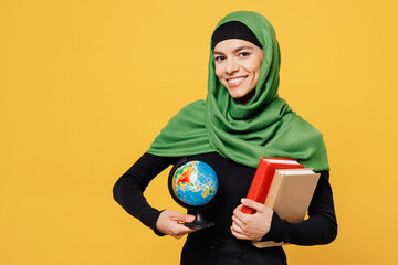 Poster - Young happy arabian asian muslim woman wear green hijab abaya black clothes hold in hand books Earth world globe isolated on plain yellow background. People uae middle eastern islam religious concept.