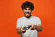 Leinwandbild Motiv Young overjoyed excited fun happy Indian man he wears t-shirt casual clothes hold in hand play pc game with joystick console isolated on orange red color background studio portrait. Lifestyle concept.