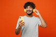 Leinwandbild Motiv Young smiling cheerful happy Indian man he wears t-shirt casual clothes listen to music in headphones use mobile cell phone isolated on orange red color background studio portrait. Lifestyle concept.