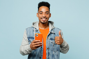 Wall Mural - Young fun man of African American ethnicity wears denim jacket orange t-shirt hold in hand use mobile cell phone show thumb up isolated on plain pastel light blue cyan background. Lifestyle concept.