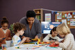 © Seventyfour - Portrait of kind Black woman helping group of little children doing crafts in preschool class, copy space