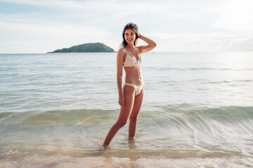 Wall Mural - Beautiful caucasian woman in white bikini on tropical beach. Portrait of dark skinned woman smiling at sea. Brunette tanned girl in swimwear enjoying and walking on beach.