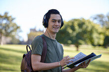 Young Man With Dark Hair Outside Holding Device Wearing Headphones At Park