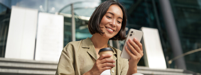 Wall Mural - Beautiful smiling asian girl, drinking coffee, using mobile phone and sits on stairs outside. Young woman video chat outdoors