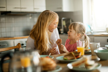 young mother embracing her daughter while having breakfast together in the kitchen at home