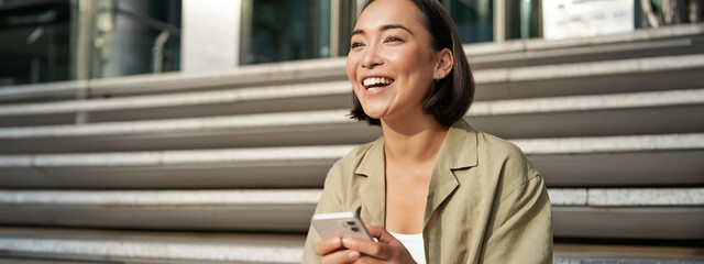 Wall Mural - Beautiful asian girl sits on stairs with smartphone. Young korean woman resting outdoors, using mobile phone application