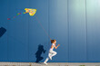 Joyful young European blonde girl in a white T-shirt and white sweatpants with pink heart-shaped glasses runs with a kite in her hand against a blue wall outside