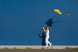 Joyful young European blond girl in a white T-shirt and white sweatpants with pink heart-shaped glasses flies a kite against a blue wall outside