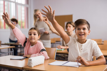 Wall Mural - Young boy and girl sitting at desk in classroom and raising hands to answer question.