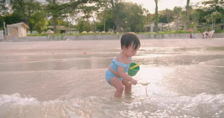 Wall Mural - Happy Cute Little Asian Toddler Child Girl in swimsuit Enjoying Vacation holding toy buckets playing sand on the beach with Waves Breaking on Seashore in summertime