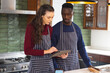 Happy diverse couple baking together in kitchen, using tablet at home