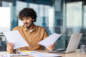 Serious and upset businessman checking and analyzing documents financial reports, thinking hispanic man reviewing contracts on paper work, man inside office at workplace with laptop.