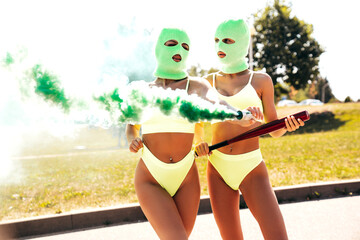 Two beautiful sexy women in green underwear. Models wearing bandit balaclava mask. Hot seductive female in nice lingerie posing in the street at sunny day. Crime and violence. Hold green smoke bomb