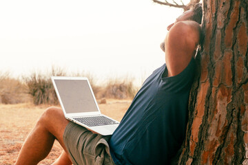 One modern traveler man after work on laptop having relax sitting at the park against a tree trunk stretching body and enjoying freedom and digital nomad smart working lifestyle. People lifestyle