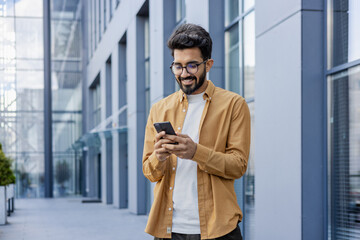 A young smiling man walks through the city with a phone in his hands, outside an office building, happily uses an application on a smartphone, reads messages, types, and browses the Internet.