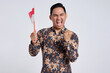 Excited young Asian man wearing batik shirt celebrate Indonesian independence day on 17 August by holding the Indonesian flag isolated over white background