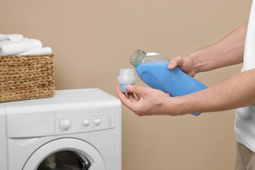 Man pouring fabric softener from bottle into cap near washing machine indoors, closeup