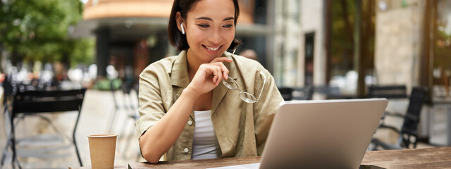 Wall Mural - Smiling young korean woman looks at her laptop screen with pleased face, works remotely from outdoors, drinks coffee