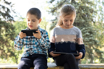 Wall Mural - Children use gadgets while sitting in the park outdoors, games on smartphone and tablet