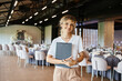© LIGHTFIELD STUDIOS - positive decorator with clipboard looking at camera near tables with festive setting in banquet hall