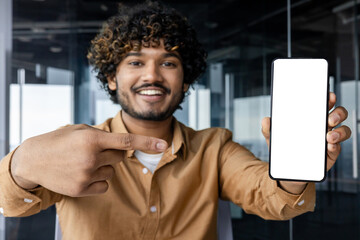 Close-up portrait of young smiling Indian man sitting in office and holding mobile phone camera, pointing finger at mockup and white mobile screen.