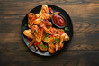 Chicken wings. Grilled or baked chicken wings with sesame seeds and ketchup or spicy tomato sauce on black plate on old wooden brawn table background. Top view with copy space.