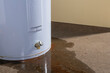 Water leaking from a residential electric water heater sitting on a concrete floor with signs of rust and iron staining the floor.