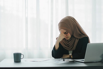 Stressed Muslim millennial businesswoman wearing a brown hijab working from home on laptop looking worried, tired and overwhelmed.