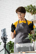 © satura_ - Relaxing home gardening. Smiling middle aged woman in black gloves with potted plant records gardening video blog in modern house - blogging and florist vlog influencer