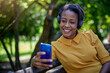 © kerkezz - Smiling young woman with headphones sitting on park bench and looking at phone.