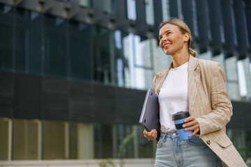 Smiling business woman drinking coffee during break time near office building and looks away 