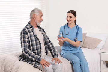 Canvas Print - Young healthcare worker giving glass of water to senior man indoors