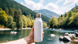 Hand holding a bottle of water in front of a mountain and a lake, hand Holding A Reusable Bottle While Hiking