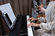 Woman In White Dress Press Key On , Tablet And Key Note Put On  Holder, And Her Friend Playing Flute Beside With Blurred Guitar And Home. Playing Music Together At Home For Fun And Relaxation.
