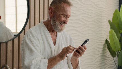 Caucasian relaxed senior old man in bathroom in bathrobe calm mature retired male in bathroom browsing mobile phone scrolling smartphone in morning check social media morning using cellphone smiling
