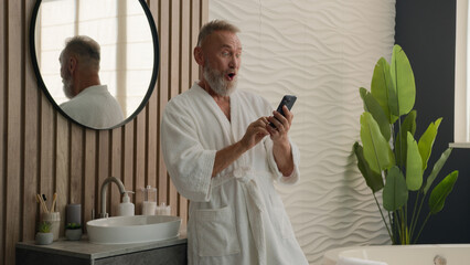 Caucasian happy old retired man in bathroom elderly senior mature male in bath morning hygiene looking mobile phone reading good news online smartphone wonder amazed surprised cellphone achievement