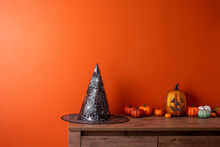Witch Hat And Pumpkins On  Wooden Chest Of Drawers Opposite  Orange Wall. Space For Text. Greeting Card Hello Autumn.
